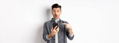 Vay canına, şuna bak. Heyecanlı yakışıklı adam telefonda teklif veriyor, akıllı telefonu işaret ediyor ve şaşkın bakıyor, beyaz arka planda duruyor..