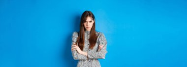 Elbiseli kızgın genç bir kadın adaletsiz hissediyor, kollarını göğsünde kavuşturuyor ve küskün, mavi arka planda duruyor..