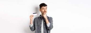 Düşünceli bir adam plastik kredi kartı tutuyor ve düşünüyor, kameraya bakıyor, seçim yaparken çenesine dokunuyor, beyaz arka planda duruyor..