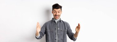 Heyecanlı beyaz adam büyük bir nesne gösteriyor, büyük bir şey şekillendiriyor ve şaşırmış bir şekilde gülümsüyor, beyaz arka planda duruyor..