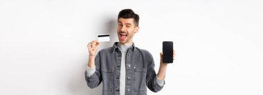 Neşeli yakışıklı adam iyi bir anlaşma yapıyor, boş akıllı telefon ekranı ve plastik kredi kartı, sana göz kırpıyor ve gülümsüyor, öneri sunuyor, beyaz arka plan..