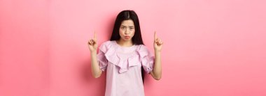 Üzgün ve karamsar Asyalı kız alnının altından bakıp somurtuyor, parmaklarını kaldırıyor ve şikayet ediyor, kıskanç ya da üzgün bakıyor, pembe arka planda duruyor..