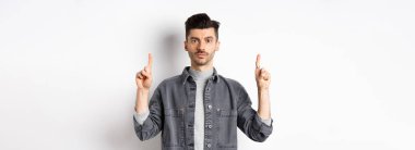 Bıyıklı, ciddi bir beyaz adam. Parmaklarını boş yere doğrultuyor ve beyaz arka planda kameraya bakıyor..