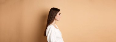 Güzel beyaz kadın profili, uzun doğal saçlı, boş alana bakan, gülümseyen romantik, bej arka planda duran..