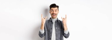 Mutlu ve heyecanlı adam jestle rock yapıyor ve eğleniyor, eğleniyor ve gülümsüyor, beyaz arka planda duruyor..