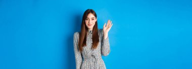 Sevimli, gülümseyen bir kadın seni selamlamak için el sallıyor, utangaç bir sırıtışla merhaba diyor, mavi arka planda giyinmiş duruyor..