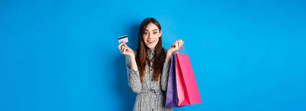 Excited beautiful female model shopping with plastic credit card, holding shop bags and smiling happy at camera, blue background.