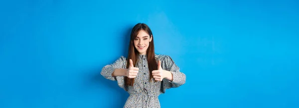Nice work, very good. Smiling young woman showing thumbs up to support your choice, looking pleased and approve something, standing in dress against blue background.