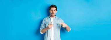 Çevrimiçi alışveriş, uygulama ve teknoloji kavramı. Etkileyici bir adam cep telefonunu işaret ediyor ve çok şaşırmış görünüyor. Uygulama öneriyor, mavi arka planda duruyor..