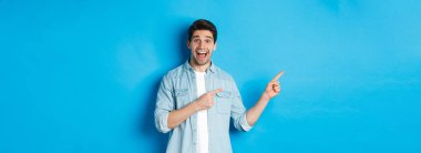 Heyecanlı yakışıklı 25 'li yaşlarda sakallı, parmaklarını sola doğru gösteren ve şaşkın bir şekilde gülümseyen, mavi arka planda duran bir adam..
