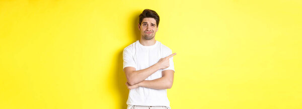 Скептически настроенный и неохотный парень в белой футболке, указывающий пальцем влево и ухмыляющийся незабавно, показывающий плохую рекламу, стоящий на желтом фоне.