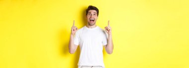 Beyaz tişörtlü neşeli genç adam tanıtım teklifine tepki veriyor, şaşkınlıkla işaret ediyor ve bakıyor, sarı arka planda duruyor..