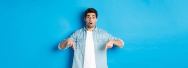 Gündelik kıyafetli sakallı, şaşırmış bir yetişkin adam, parmakla işaret ediyor ve şaşırmış görünüyor, terfi hakkında sorular soruyor, mavi arka planda duruyor..