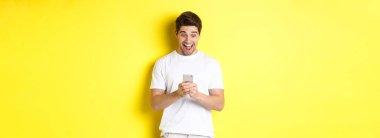 Şaşırmış bir adam cep telefonundan mesaj okuyor, akıllı telefon ekranında şaşırmış ve mutlu görünüyor, sarı arka planda duruyor..