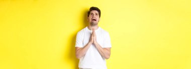 Çaresiz bir adam Tanrı 'ya yalvarıyor, el ele tutuşup dua ediyor, kederli bir şekilde yukarı bakıyor, sarı arka planda dikiliyor. Boşluğu kopyala