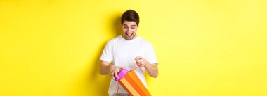 Tatil ve kutlama kavramı. Sarı arka planda duran genç adam, alışveriş çantasından hediye alırken şaşırmış görünüyordu..