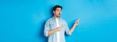 Şaşırmış ve şaşırmış bir adam terfiye bakıyor, parmaklarını reklamlara gösteriyor, mavi arka planda duruyor..