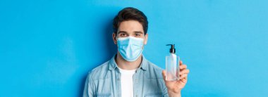 Koronavirüs kavramı, pandemik ve önleyici tedbirler. Tıbbi maske takan genç adama el dezenfektanı kullanmasını ve mavi arka planda duran antiseptik şişeyi göstermesini tavsiye ediyoruz..