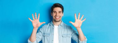 Yakından bakınca, gülümseyen, on numarayı gösteren, mavi arka planda duran yakışıklı bir adam..