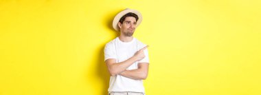 Turizm ve yaşam tarzı kavramı. Memnun olmayan erkek turist şikayetçi, sol üst köşedeki tanıtıma bakıyor ve işaret ediyor, hayal kırıklığıyla, sarı arka planda duruyor..