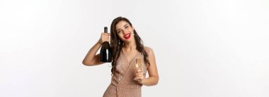 Kış bayramı kutlaması. Mutlu bir kadın Noel partisinde kadeh kaldırıyor, şampanya şişesini kaldırıyor ve gülümsüyor, zarif bir elbise içinde beyaz arka planda duruyor..