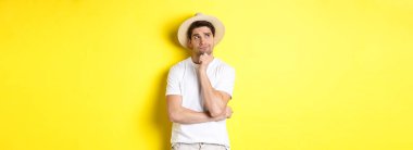 Turizm ve yaz kavramı. Düşünceli bir turist, sol üst köşeye bakıyor ve düşünüyor, saman şapka ve beyaz tişörtle sarı arka planda duruyor..