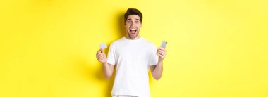 Elinde akıllı telefon ve kredi kartı olan şaşırmış bir adam. Kara Cuma 'da online alışveriş yapıyor. Sarı arka planda duruyor..