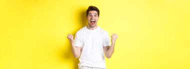 Heyecanlı ve şanslı adam kazanıyor, yumruklarını sıkıyor ve mutlu görünüyor, zafer kazanıyor ve kutluyor, sarı arka planda duruyor..