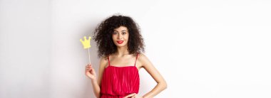 Kırmızı elbiseli şık bir genç kadın, kendinden emin ve küstah, tacını tutuyor ve gülümsüyor, beyaz arka planda duruyor..