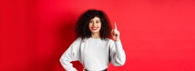 Bir numarayı gösteren kıvırcık saçlı modern Avrupalı kadın portresi, sipariş veriyor, parmağını kaldırıyor ve gülümsüyor, kırmızı arka planda duruyor..