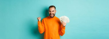 Alışverişe. Neşeli adam elinde para tutuyor, ödül kazanıyor ve yumruk tokuşturuyor, tatmin olmuş bir ifadeyle zafer kazanıyor turkuaz arka planda turuncu süveterin içinde duruyor..