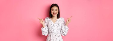Güzel Asyalı kadın alışverişte seçim yapıyor, parmaklarını yana doğru gösteriyor ve değişkenleri gösteriyor, kameraya gülümsüyor, pembe arka planda duruyor..