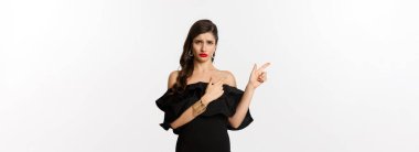 Moda ve güzellik. Hayal kırıklığına uğramış cazibeli kadın şikayet ediyor, siyah elbise giyiyor, somurtuyor ve kötü teklifi işaret ediyor, beyaz arka plan.