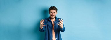 Öfkeli adam birini boğmak ister, düşmanını boğmak için ellerini uzatır, dudaklarını ısırır ve öfkeli görünür, mavi arka planda durur..