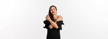 Moda ve güzellik. Siyah elbiseli, şehvetli ve güzel bir kadın, kapalı gözlerle kendine sarılıyor, hayal kuruyor, beyaz arka planda duruyor..