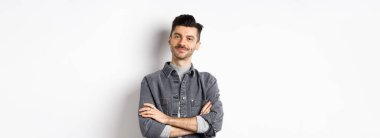 Gururlu yakışıklı adam tatmin olmuş görünüyor, göğsünde çapraz kollar ve kameraya gülümsüyor, iyi bir şeye bakıyor, beyaz arka planda duruyor..