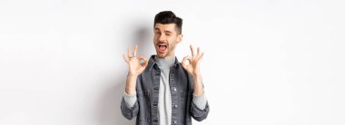 Her şey yolunda. Yakışıklı beyaz adam iyi işaretler gösteriyor ve gülümsüyor, iyi şeyi övmek için göz kırpıyor, mükemmel bir teklif sunuyor, beyaz arka planda duruyor..