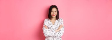 Güzel Asyalı kadın model elbise giymiş, kolları göğsünde ve kameraya gülümsüyor, pembe arka planda duruyor..