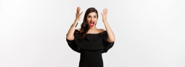 Moda ve güzellik. Siyah elbiseli kızgın bir kadın, deli gibi bağırıyor ve el sıkışıyor, kamera karşısında suratını ekşitiyor, beyaz arka planda duruyor..