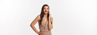 Noel partisi ve kutlama konsepti. Kırmızı dudaklı çekici genç bir kadın, lüks elbiseler giyiyor, cep telefonuyla konuşuyor ve gülümsüyor, beyaz arka planda duruyor..