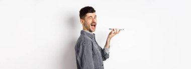 Mutlu adam kameraya bakarken ses çevirici uygulamasını kullanıyor ya da hoparlörden konuşuyor, telefonu ağıza yakın tutuyor, beyaz arka planda duruyor..