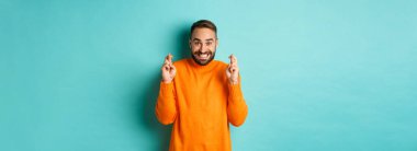 Sakallı heyecanlı bir adam, bir dilek diliyor, şans diliyor ve gülümsüyor, açık mavi arka planda duruyor..