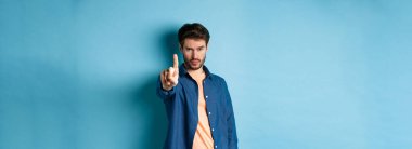 Ciddi genç adam seni durdurmak ya da uyarmak için bir parmağını uzatıyor, bir şeyi yasaklıyor ya da onaylamıyor, mavi arka planda duruyor. Boşluğu kopyala