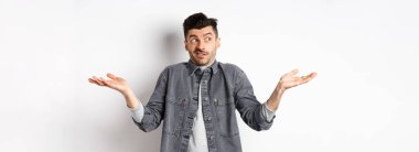 Kararsız genç adam yana bakıyor ve omuz silkiyor, hiçbir şey bilmiyor, hiçbir fikri yok, beyaz arka plana karşı cahil ve kafası karışık duruyor..