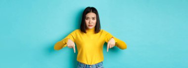 Alışveriş konsepti. Üzgün ve endişeli Asyalı kız, aptal gibi kaşlarını çatıyor, hatayı işaret ediyor, mavi arka planda üzgün duruyor..