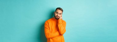 Üzgün ve kasvetli sakallı bir adam, kararsız ve üzgün görünüyor, bir şeyler düşünüyor ve somurtuyor, turkuaz arka planda duruyor..