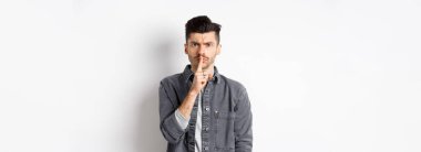 - Sessiz ol. Kızgın adam somurtuyor ve susuyor, sakin olmasını söylüyor, kötü davranışları azarlamak için parmaklarını dudaklarına bastırıyor, beyaz arka planda duruyor..