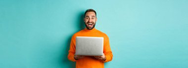 Mutlu adam dizüstü bilgisayar kullanıyor ve kameraya heyecanlı bakıyor, açık mavi arka planda bilgisayarla duruyor..
