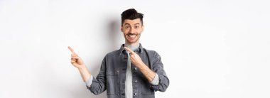 Bıyıklı, beyaz gülüşlü, sağı gösteren, buraya bakmaya davet eden, beyaz arka planda duran neşeli, şık bir adam..