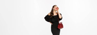 Güzellik ve moda konsepti. Siyah elbise, topuklu ayakkabı ve çantayla alışverişe çıkan, kendine güvenen, beyaz arka planda duran zarif genç bir kadın..